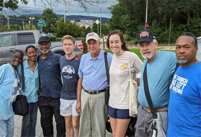 Eight people in a line with arms around each other shoulders smile for the camera. In the distance a gloomy cloud threatens rain.